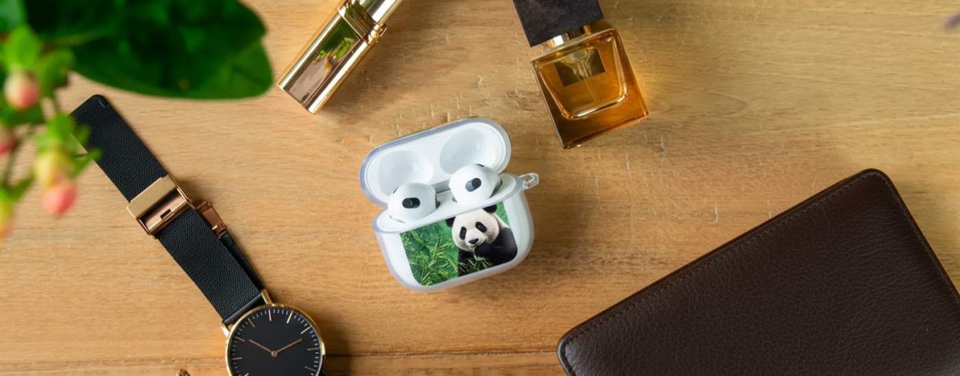 An overhead view of an open white earbud charging case featuring a photographic print of a giant panda eating bamboo, placed on a wooden desk alongside a black watch, a leather wallet, and a perfume bottle.