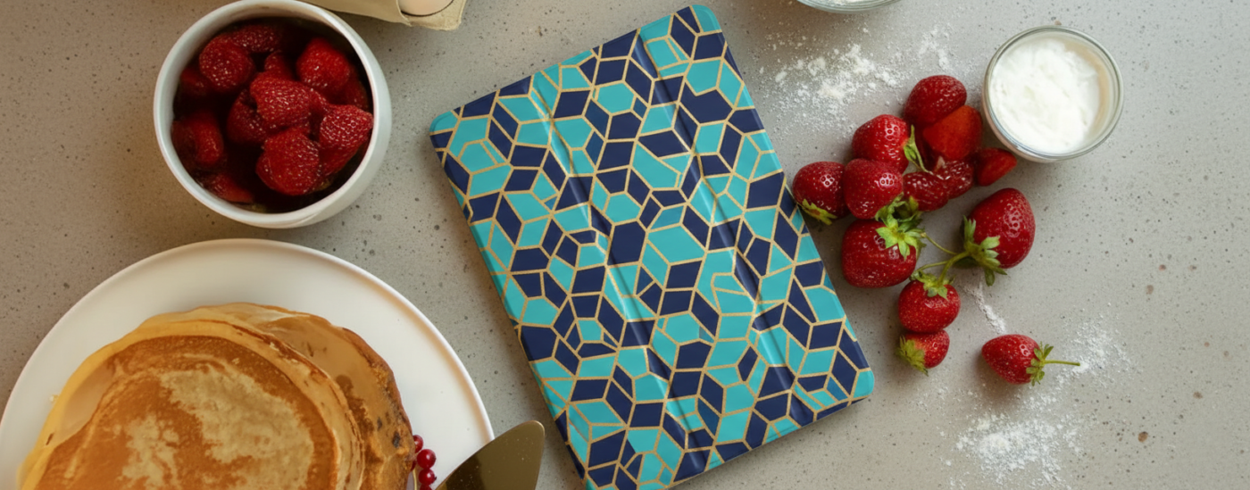 A top-down view of a modern teal and navy geometric patterned tablet cover lying on a stone countertop next to a plate of pancakes, fresh strawberries, and bowls of yogurt and berry compote